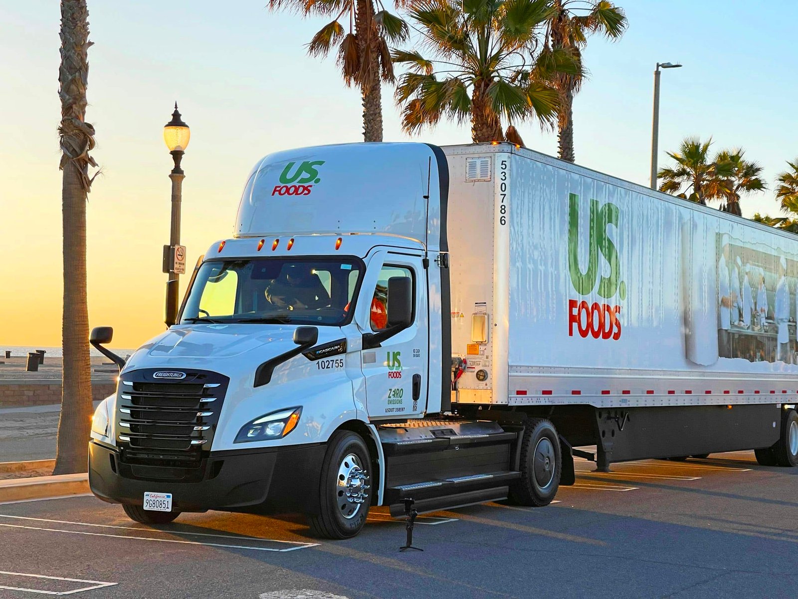 A US Foods semi-trailer truck parked by the beachside in Huntington Beach, CA during sunset with palm trees and street lamps lining the view.